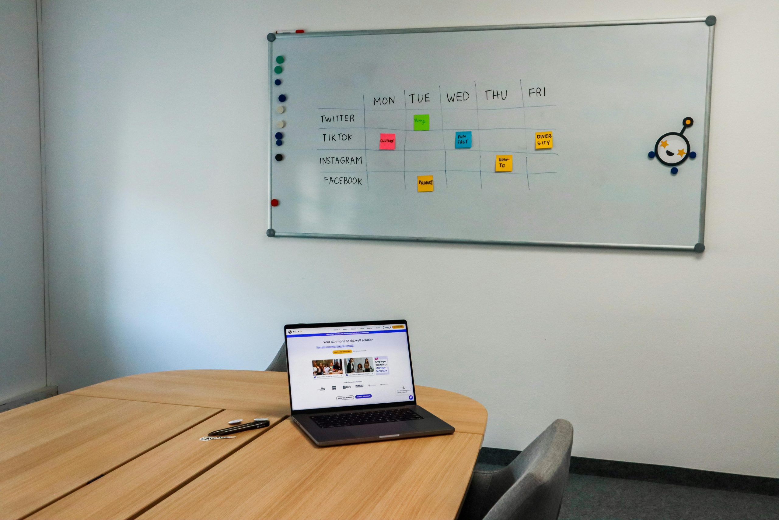Laptop on a meeting table beneath a whiteboard mapping a weekly posting plan across social channels